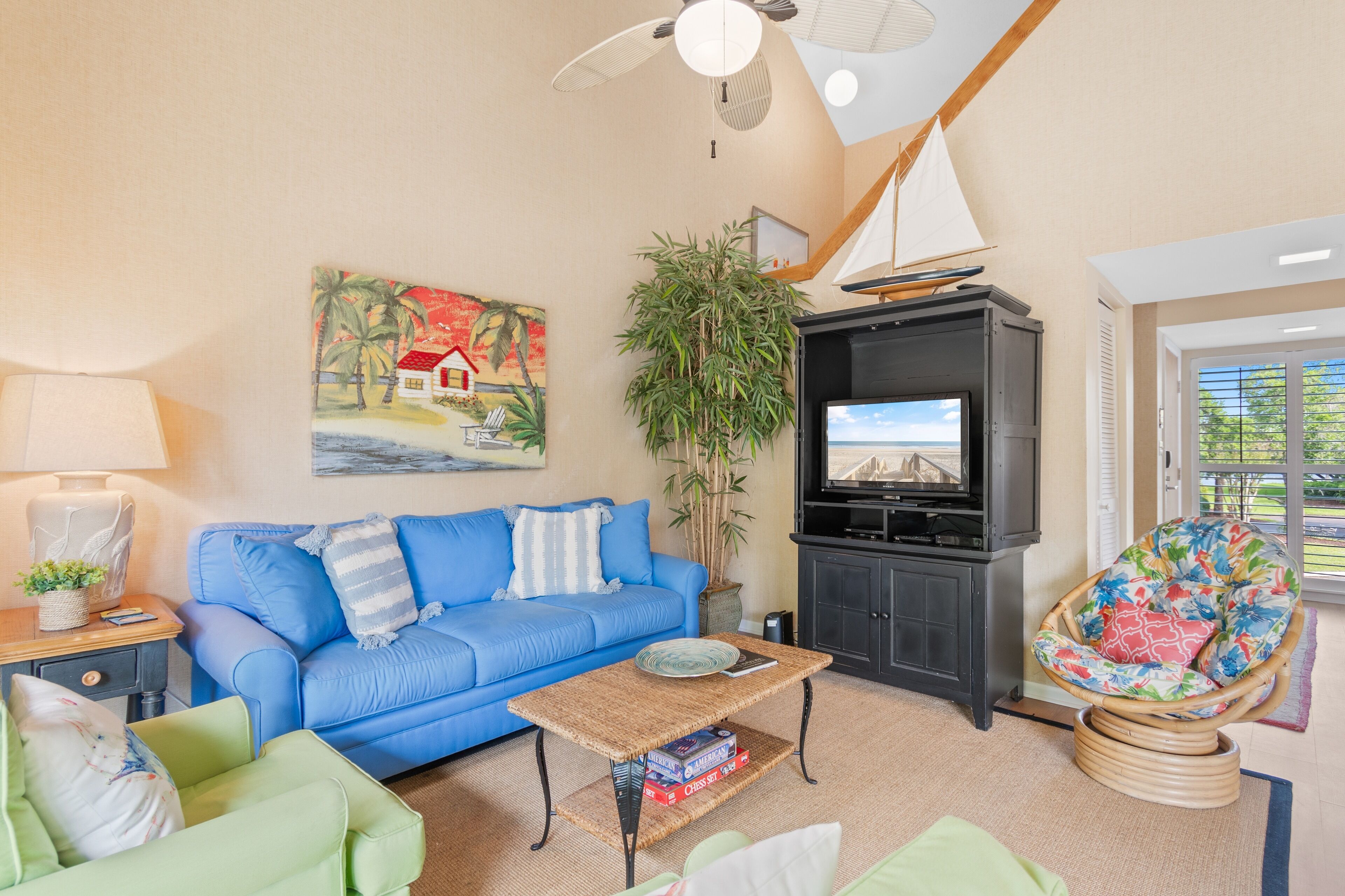 A living room with a blue sofa, a green armchair, a wicker chair, a television, a bookshelf, and a painting of a beach scene.