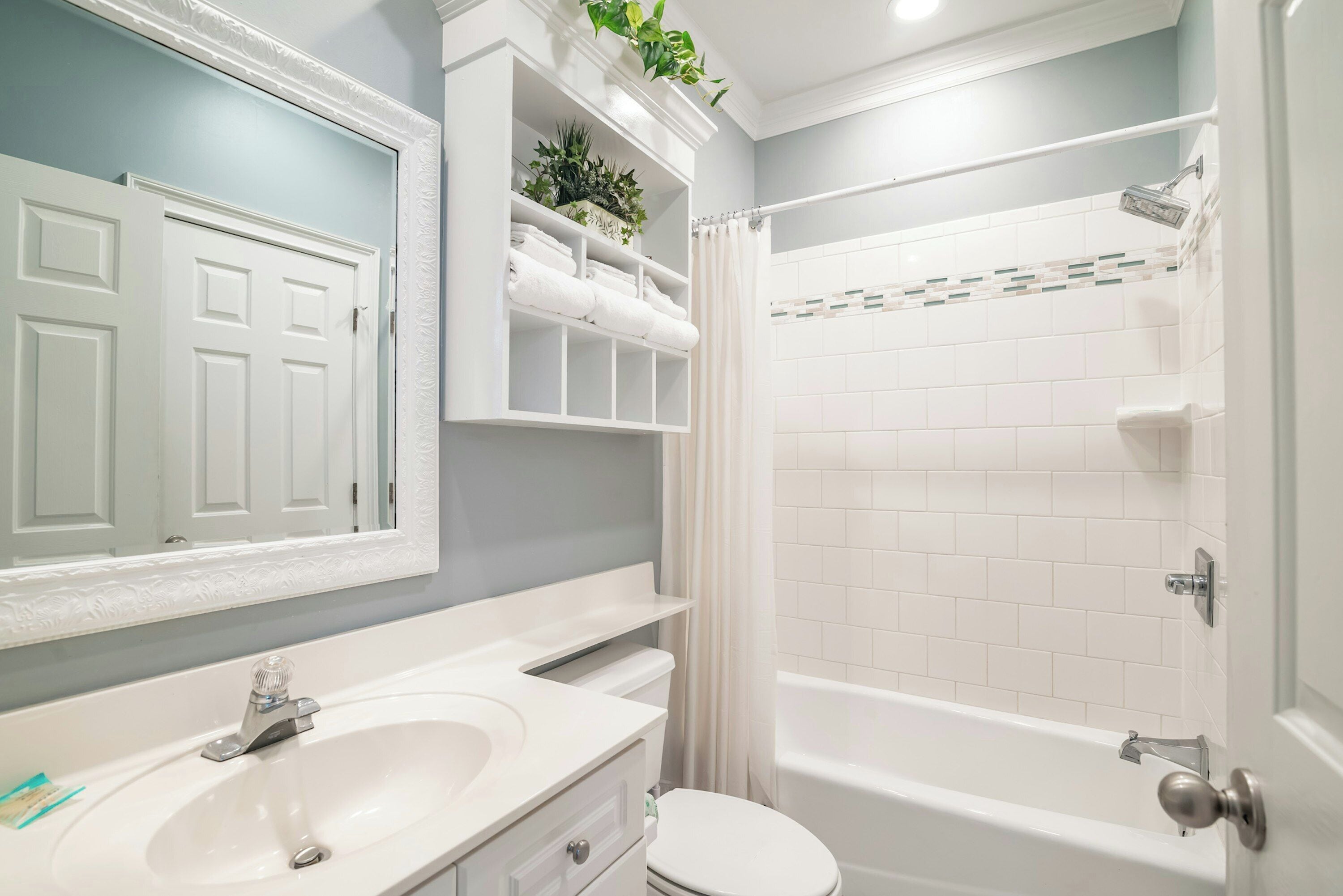 Une salle de bain avec un lavabo, des toilettes et une baignoire, tous de couleur blanche. Un miroir est placé au-dessus du lavabo, et une étagère contient des serviettes et des plantes.