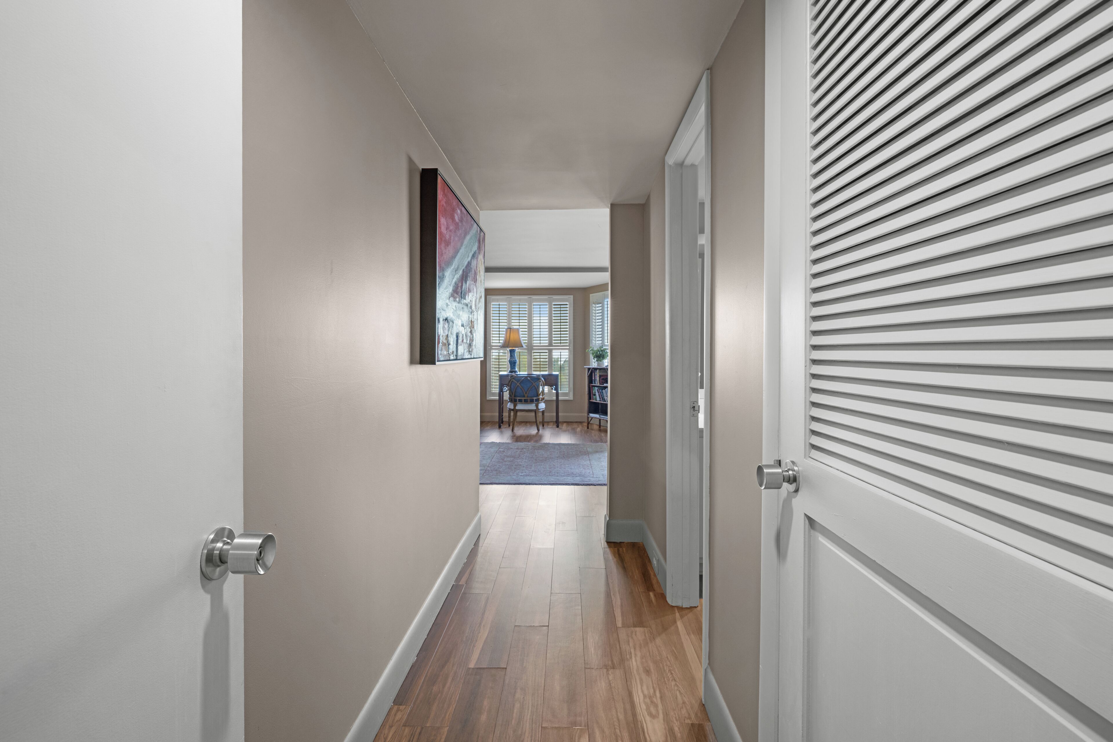 Un couloir avec un plancher en bois, une porte dotée d’un cadre blanc et un tableau accroché au mur.