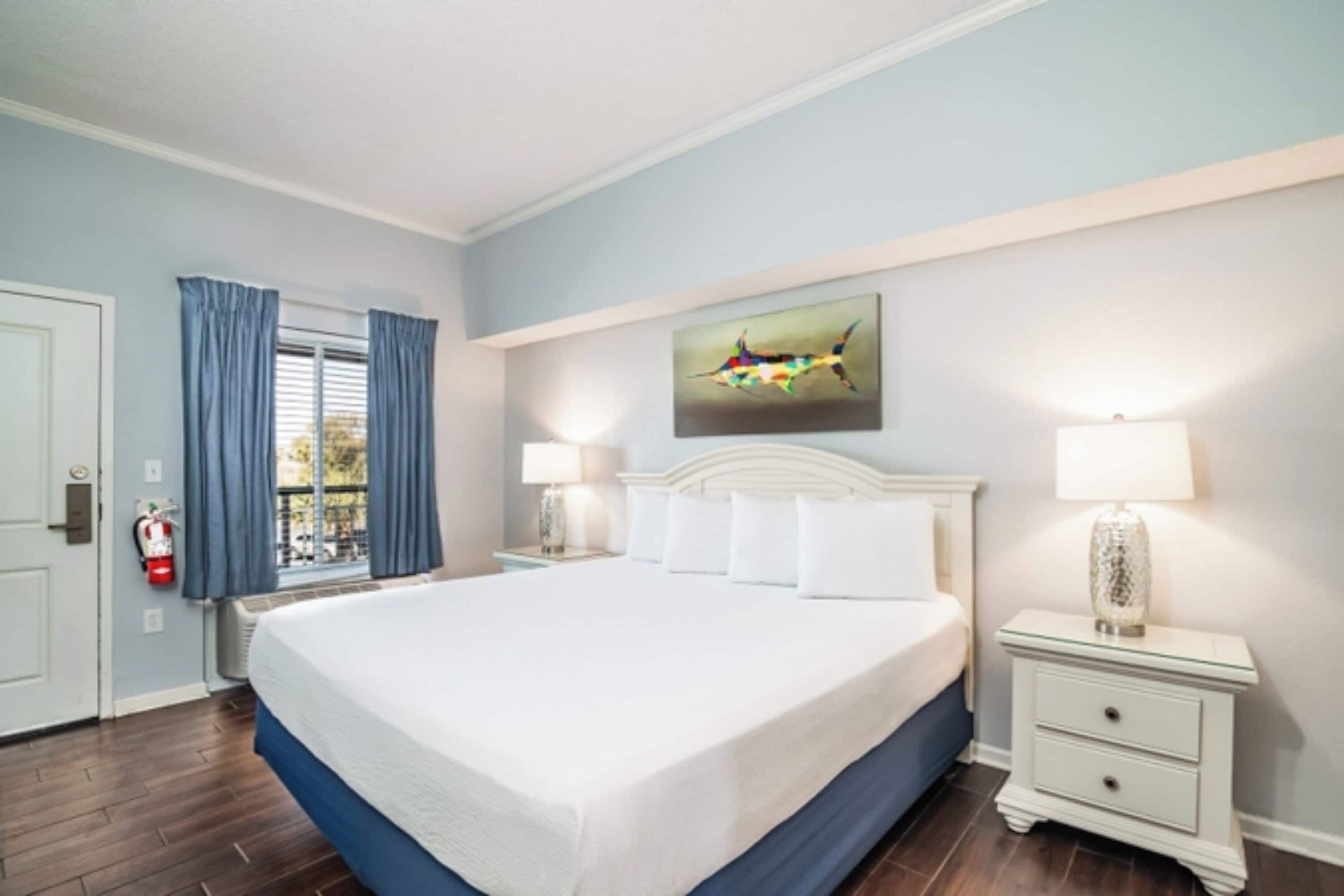 Een hotelkamer met een groot bed, twee nachtkastjes met lampen, een raam met blauwe gordijnen, een brandblusser en een wanddecoratie.