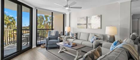 A spacious living room with a large window, a ceiling fan, and a balcony with outdoor furniture.
