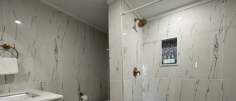 A modern bathroom with a marble wall, a shower with a gold showerhead, a toilet, and a sink with a towel.