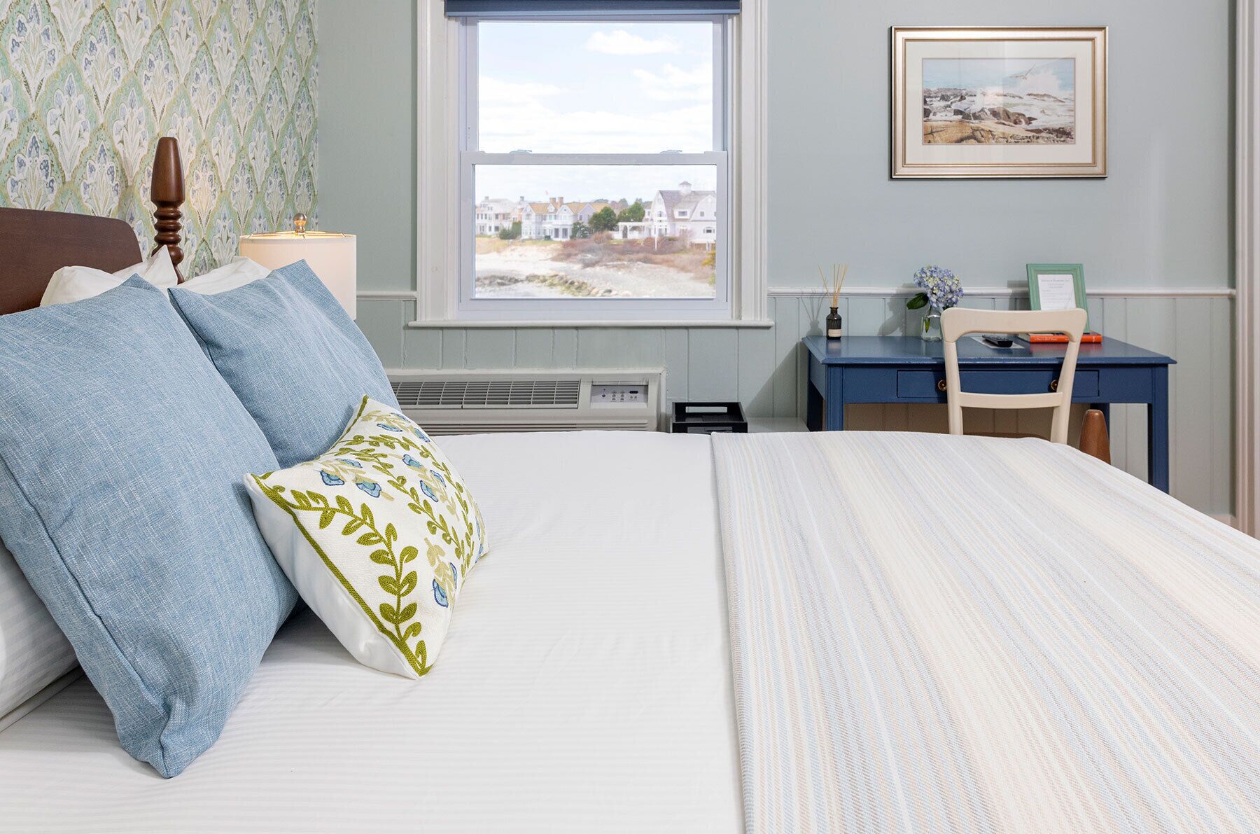 A bedroom with a bed, a desk, a chair, a window with a view of houses, and a framed picture on the wall.