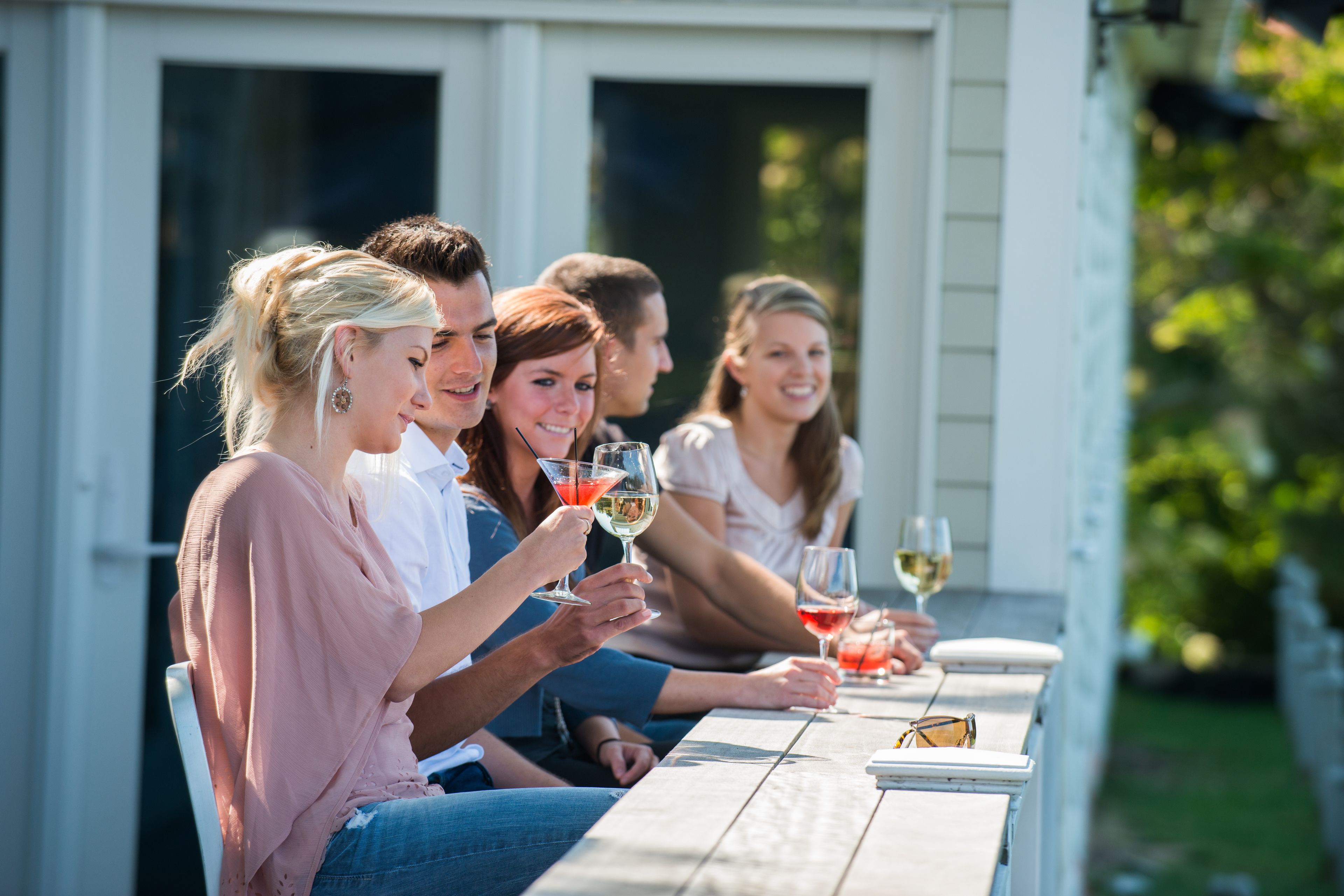 Un groupe de personnes est assis à une table à l’extérieur, devant des boissons. L’endroit se caractérise par une façade blanche et un décor verdoyant en arrière-plan.