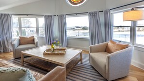 Ein modernes Wohnzimmer mit Sofa, Sessel und Couchtisch, großen Fenstern mit Vorhängen und Blick ins Freie.