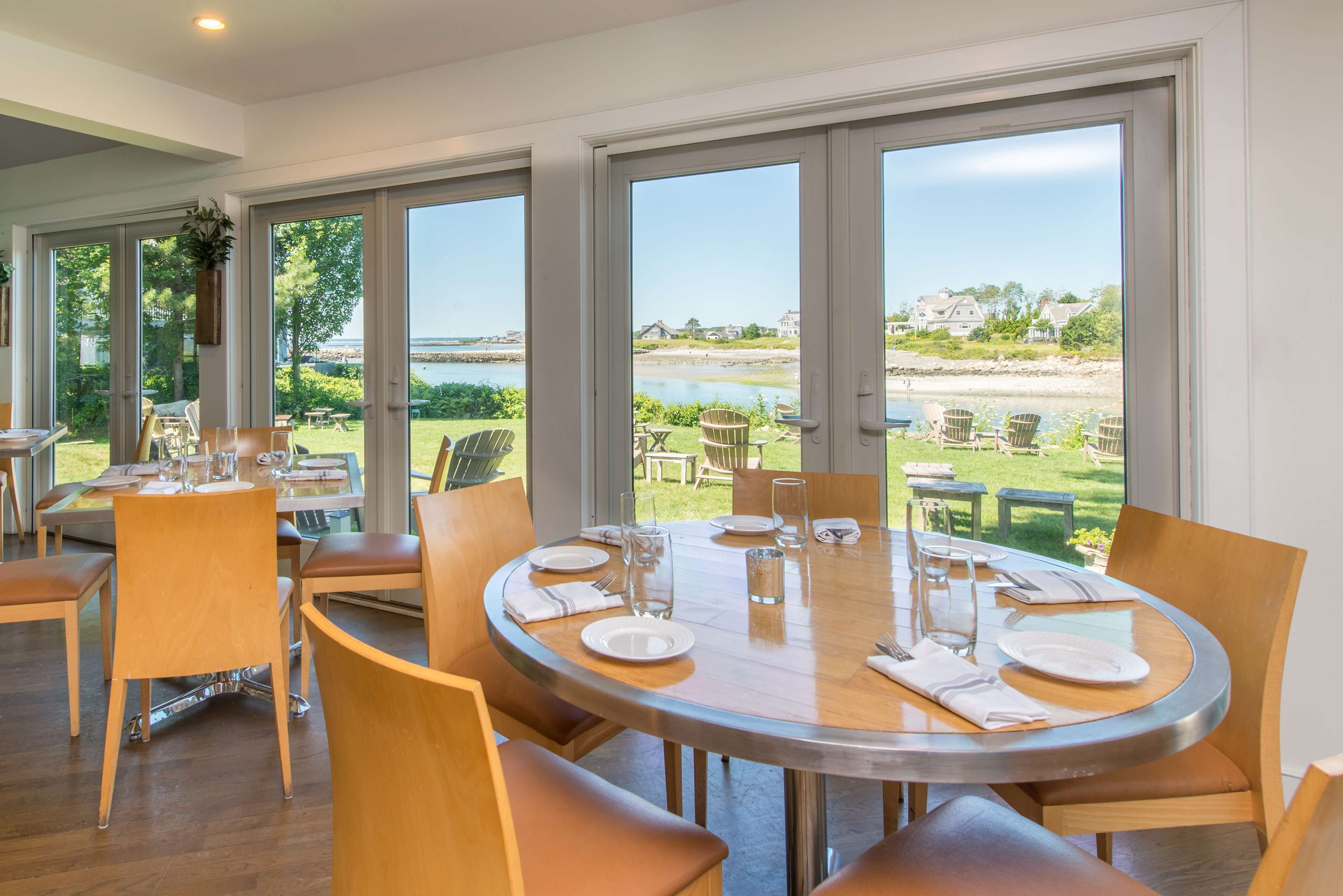 A dining area with a round table set for six, large windows offering a view of a beach and greenery.