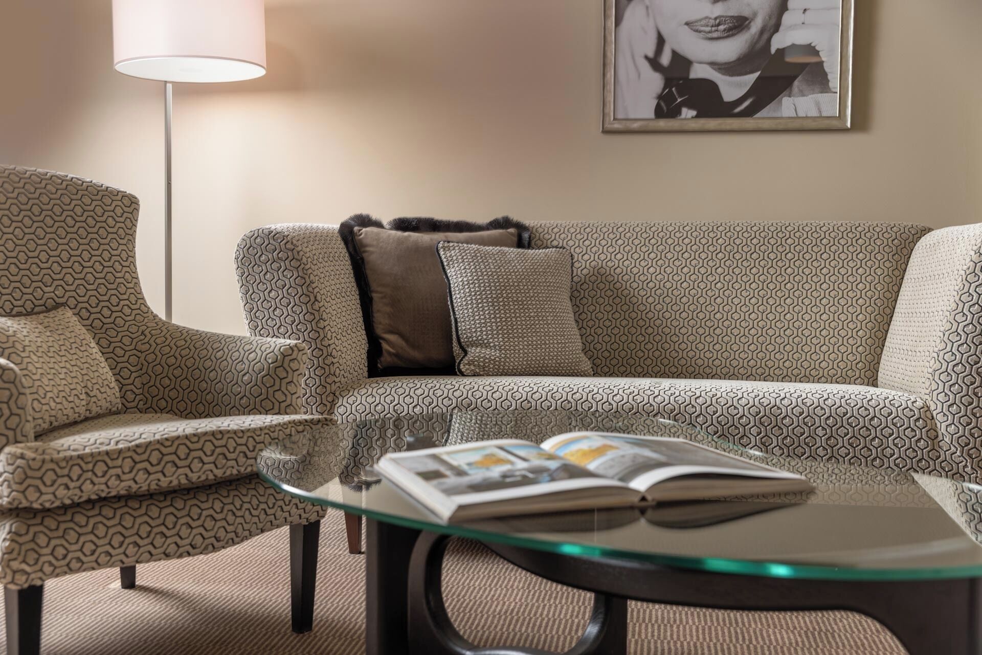 A living room with a patterned sofa, a glass coffee table with magazines, a floor lamp, and a framed picture on the wall.