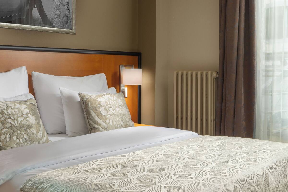 A hotel room with a large bed, a headboard, a nightstand with a lamp, a radiator, and a window with curtains.
