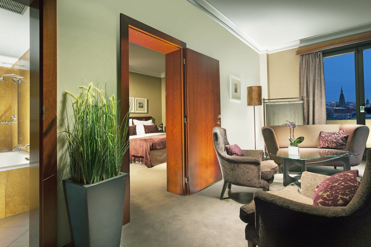 A hotel room with a bed, a sitting area with a sofa and armchairs, a glass coffee table, a potted plant, and a view of a cityscape through a window.