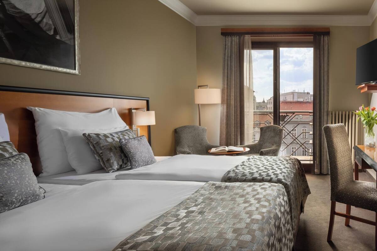 A hotel room with two beds, a desk with a lamp, a chair, and a view of the city through large windows.