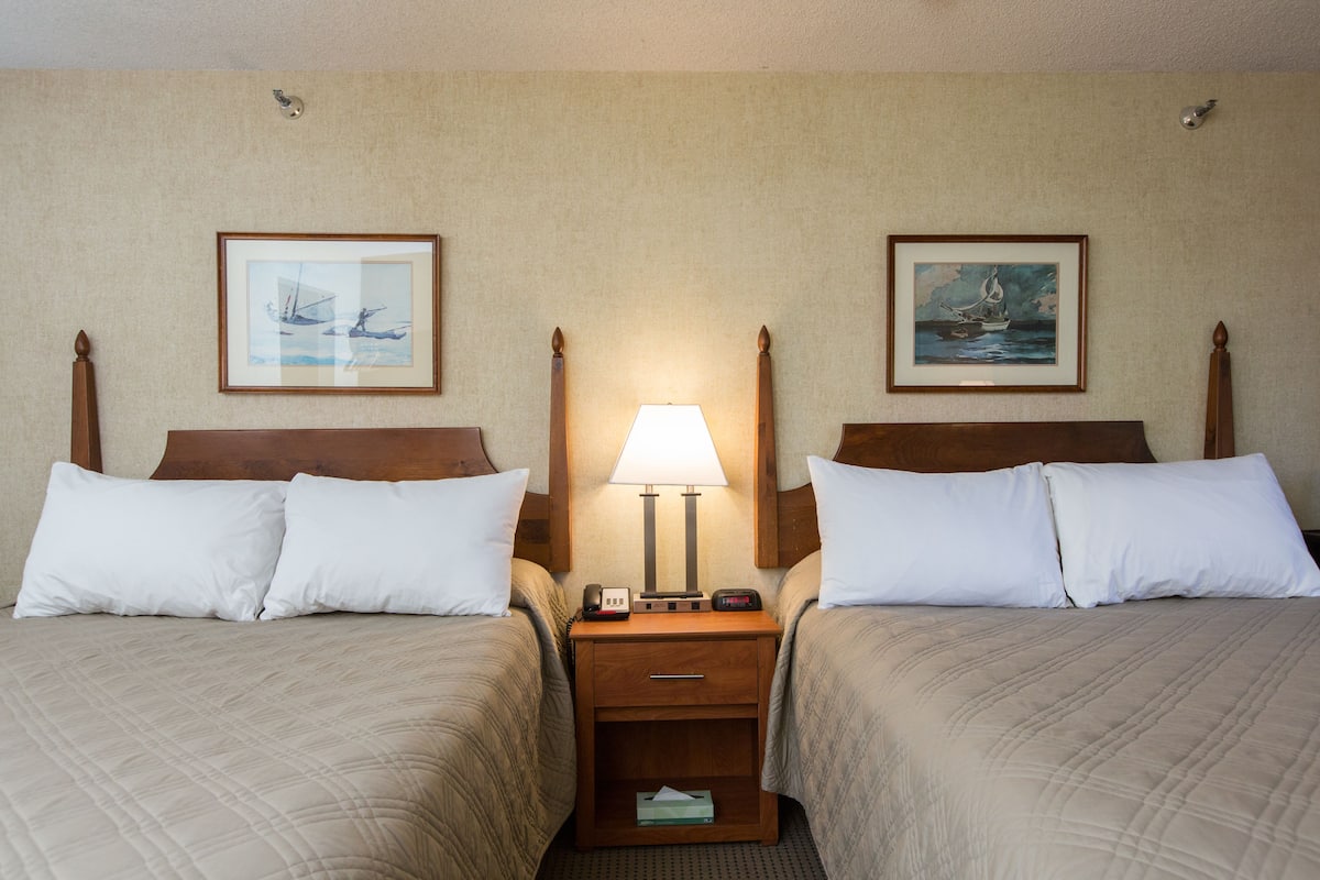 A hotel room with two beds, a nightstand with a lamp, and framed artwork on the walls.