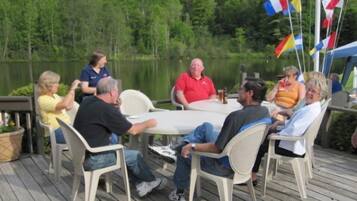 Eine Gruppe von Personen sitzt an einem Tisch auf einer Holzterrasse an einem See, im Hintergrund wehen Flaggen.