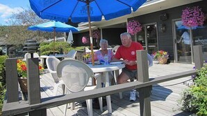 Ein Außenbereich mit einer Holzterrasse, weißen Plastikstühlen und blauen Sonnenschirmen.