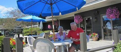 An outdoor patio area with wooden deck, white plastic chairs, and blue umbrellas.