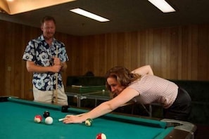A pool table with balls and a cue stick, a person leaning over the table, and another person standing beside it.