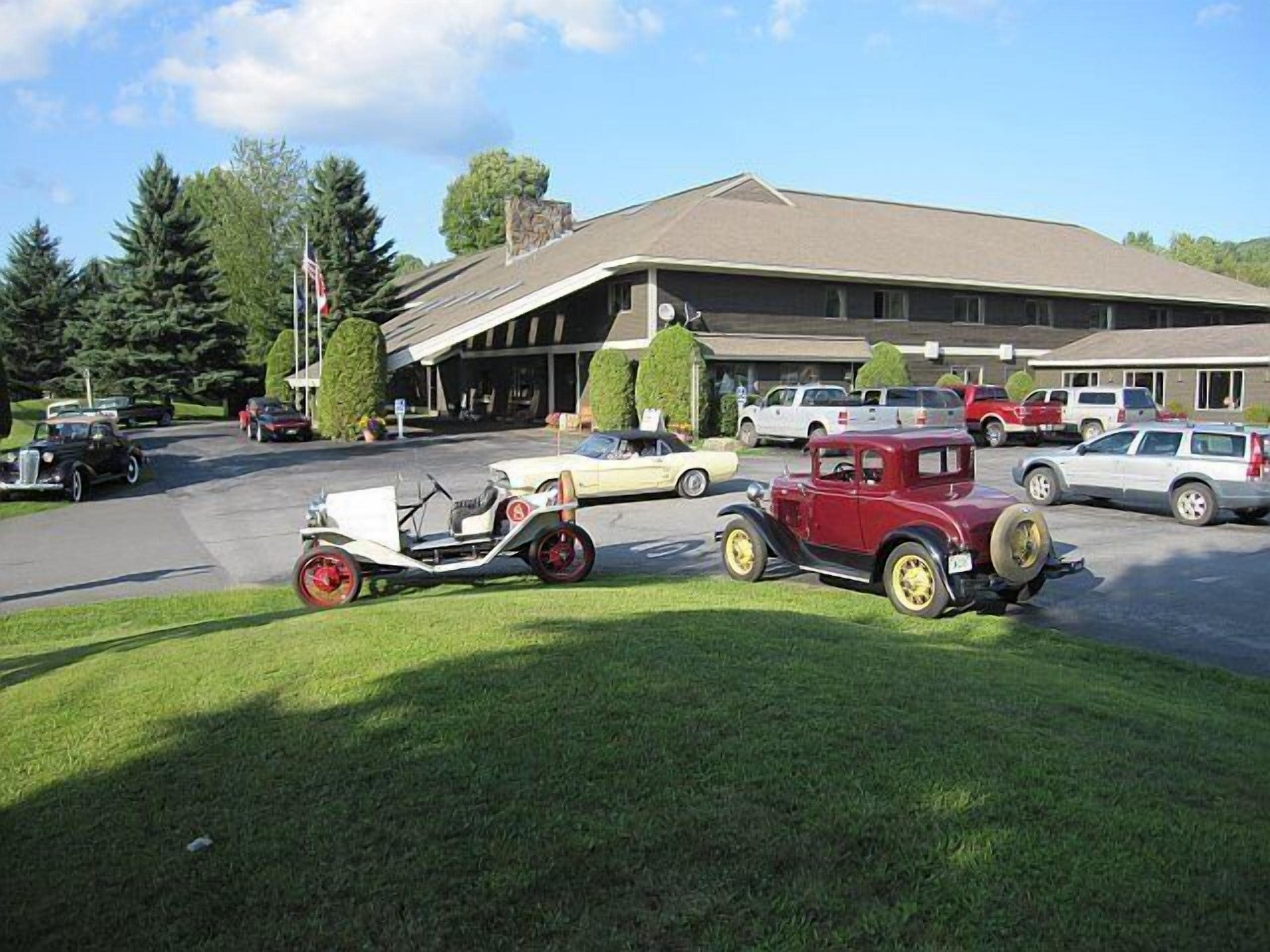 A parking lot with several vintage cars, a building with a flat roof, and a grassy area in the foreground.