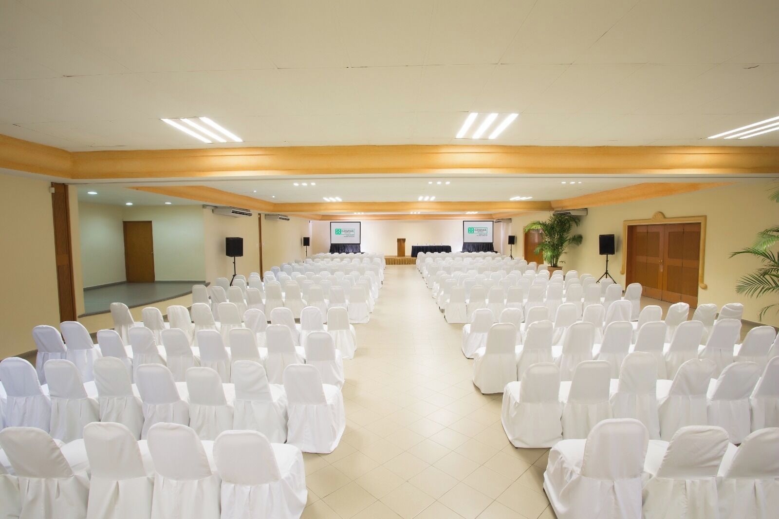 A conference room with rows of white chairs, a stage with a screen, and a door on the left.