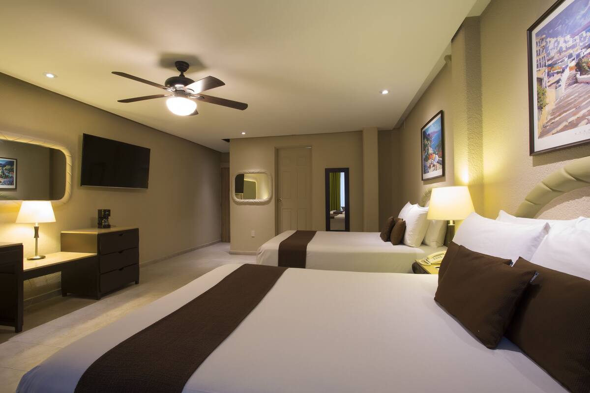 A hotel room with two beds, a ceiling fan, a desk with a lamp, a dresser, and a TV on the wall.