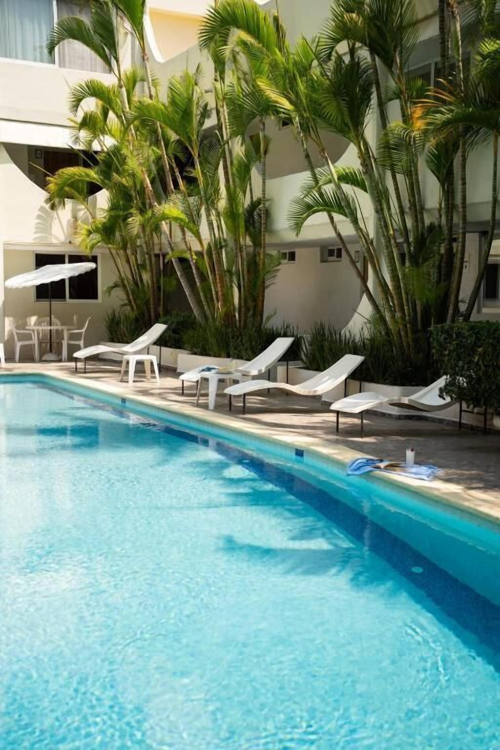 Una piscina con tumbonas, palmeras y un edificio al fondo.