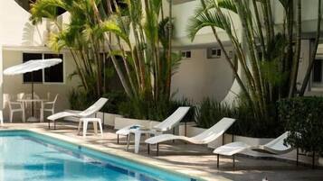 Una piscina con tumbonas, palmeras y un edificio al fondo.