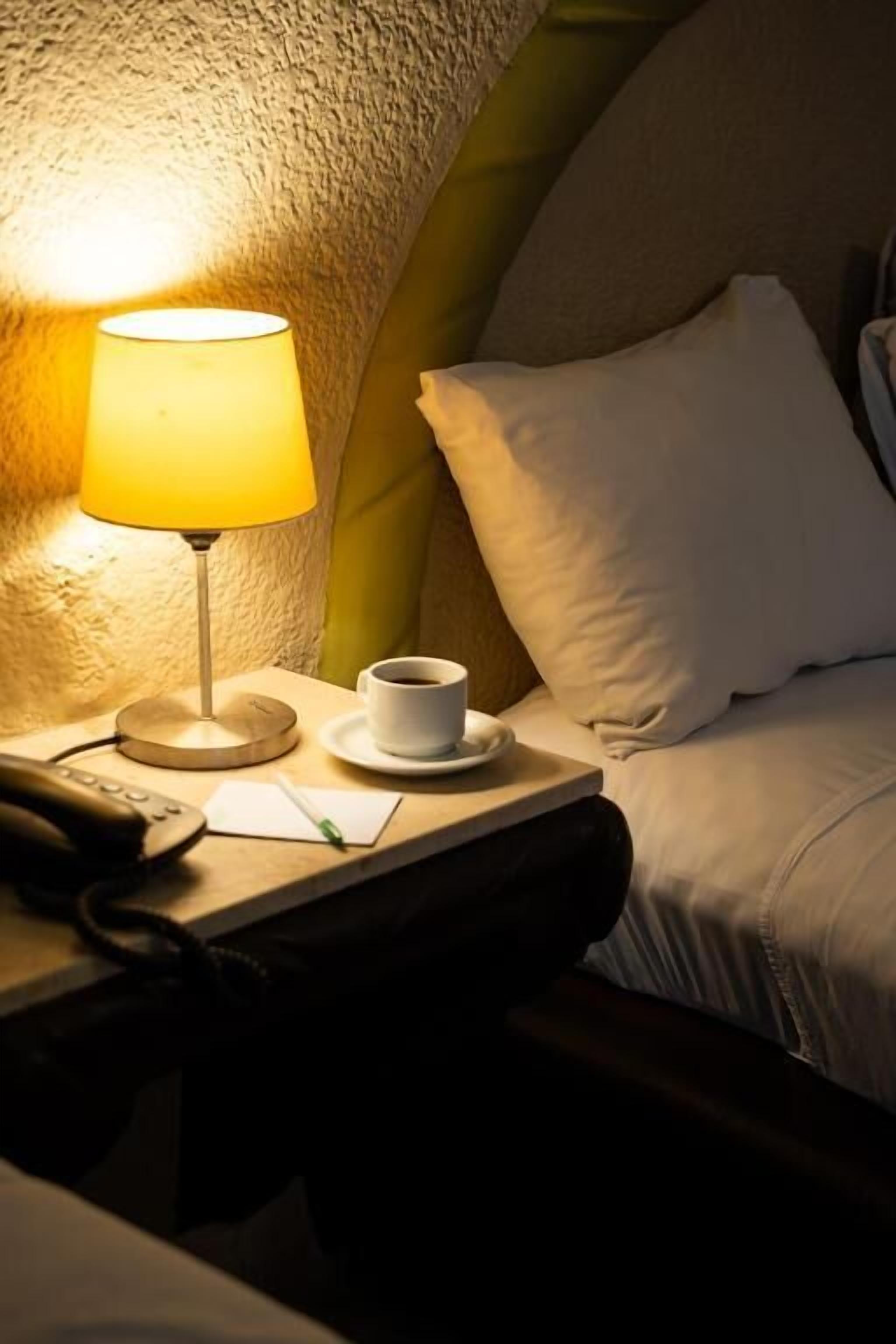 Una habitación de hotel con una lámpara de noche, una taza de café, un bloc de notas y un teléfono en la mesita de noche.