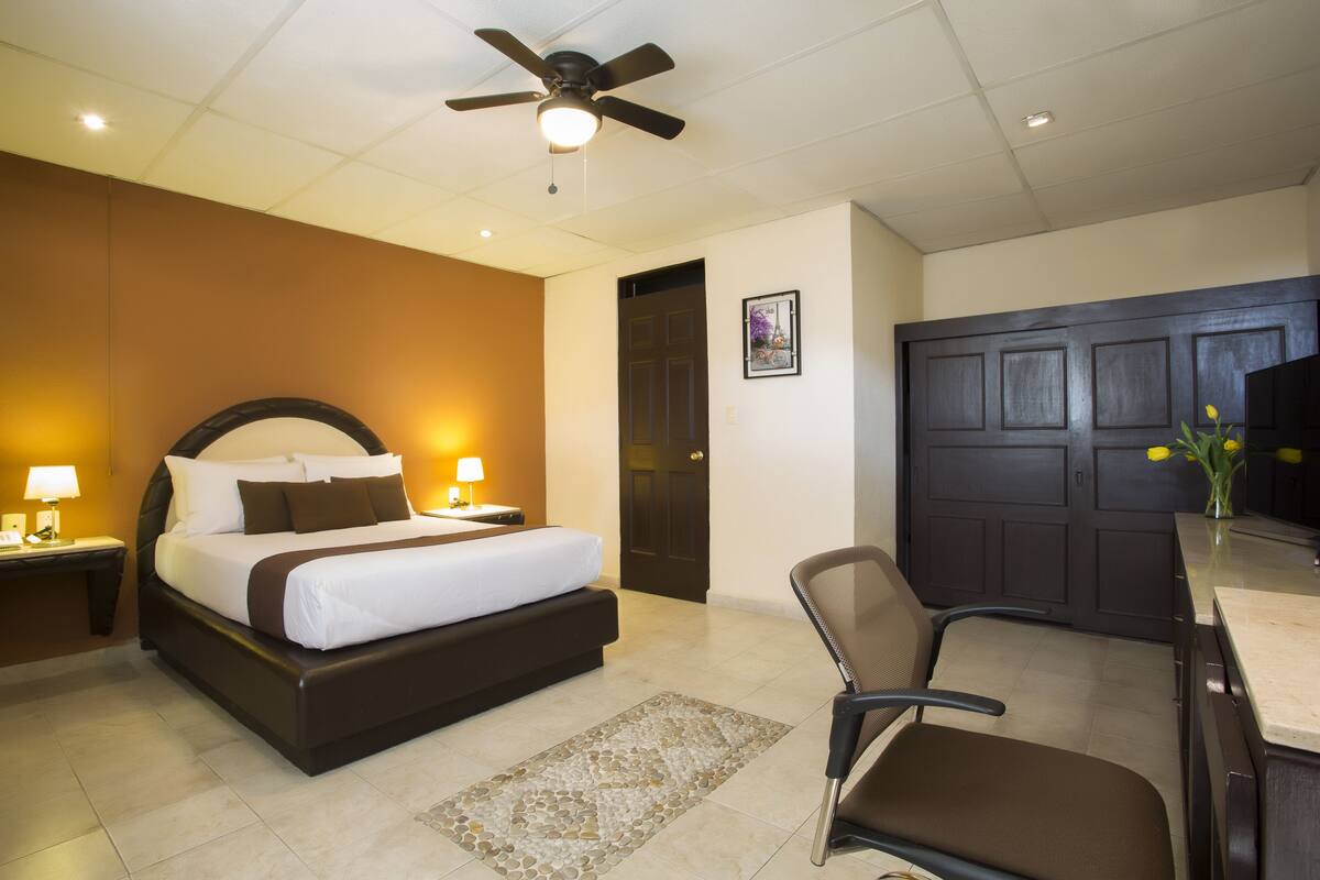 A hotel room with a bed, a desk, a chair, a ceiling fan, and a door.