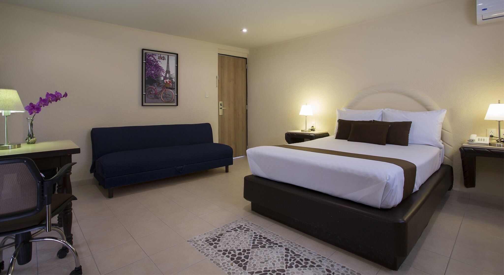 Quarto de hotel com cama, escrivaninha com luminária, cadeira, sofá e quadro na parede.