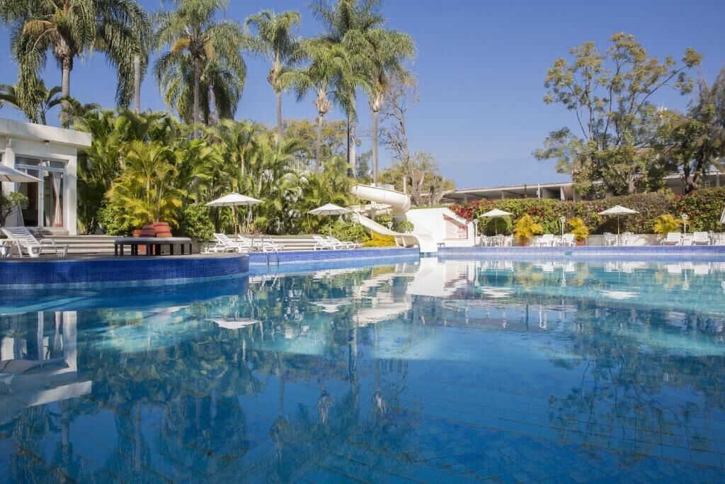 A clear blue swimming pool with lounge chairs and umbrellas, surrounded by palm trees and a modern building.