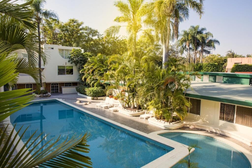 A swimming pool surrounded by palm trees and a white building with a green roof.