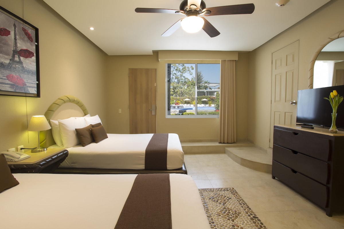A hotel room with two beds, a television, a desk with a lamp, and a view of the outdoors.