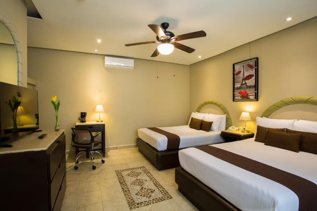 A hotel room with two beds, a desk, a chair, a ceiling fan, and a painting on the wall.