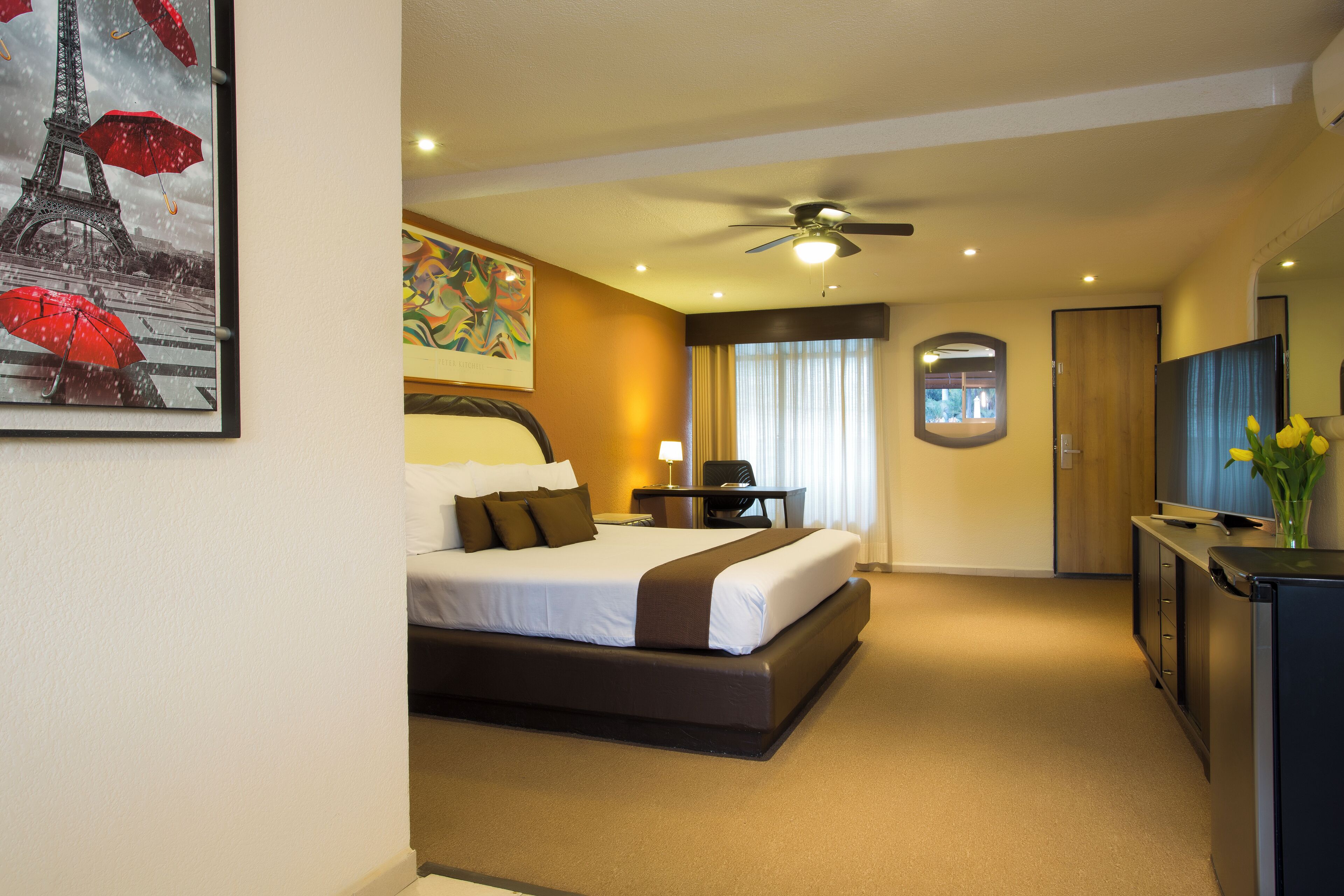 Una habitación de hotel con cama, escritorio, ventilador de techo, televisión y un cuadro de la Torre Eiffel.