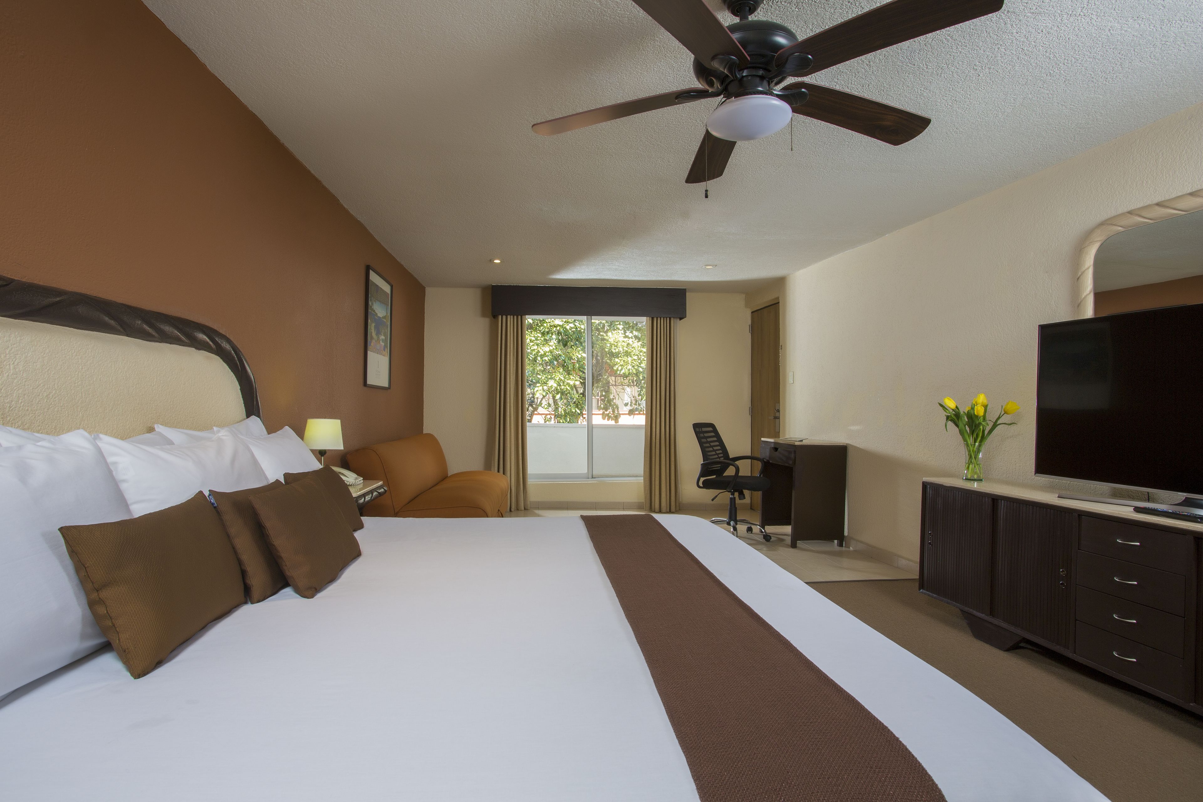 A hotel room with a large bed, a desk, a television, and a ceiling fan.