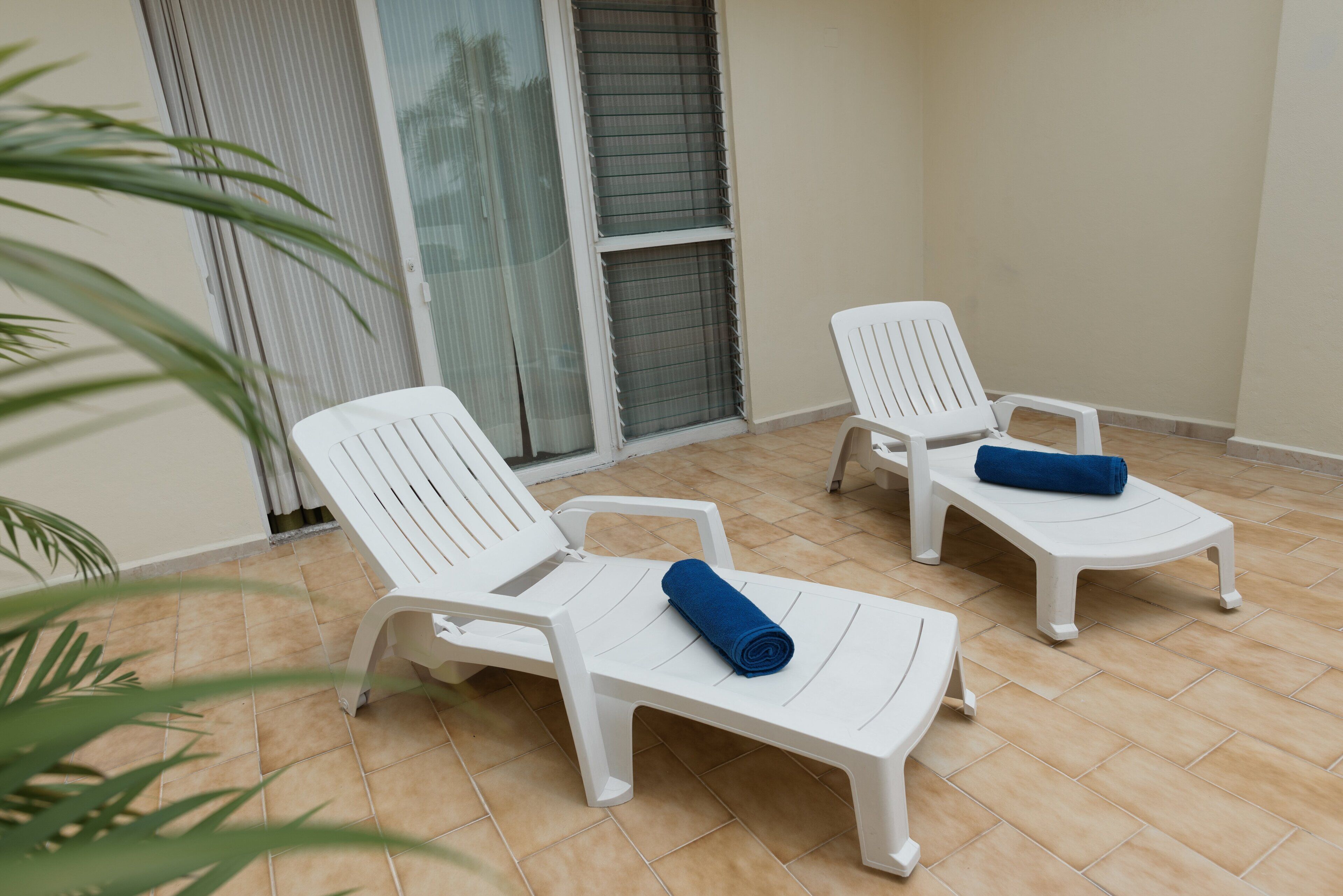 Un balcon avec deux chaises longues en plastique blanc, chacune agrémentée d’une serviette bleue roulée.