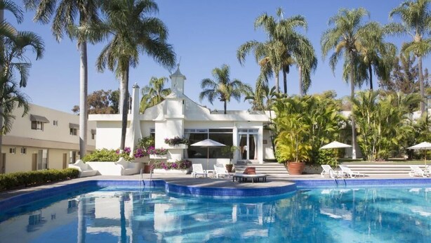 A swimming pool with lounge chairs and umbrellas, surrounded by palm trees and a building with a white facade. - Gamma Cuernavaca Puerta Paraiso (Cuernavaca)