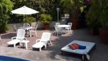 Zona de piscina con sillones de descanso y una mesa, rodeada de plantas en macetas y un edificio al fondo.