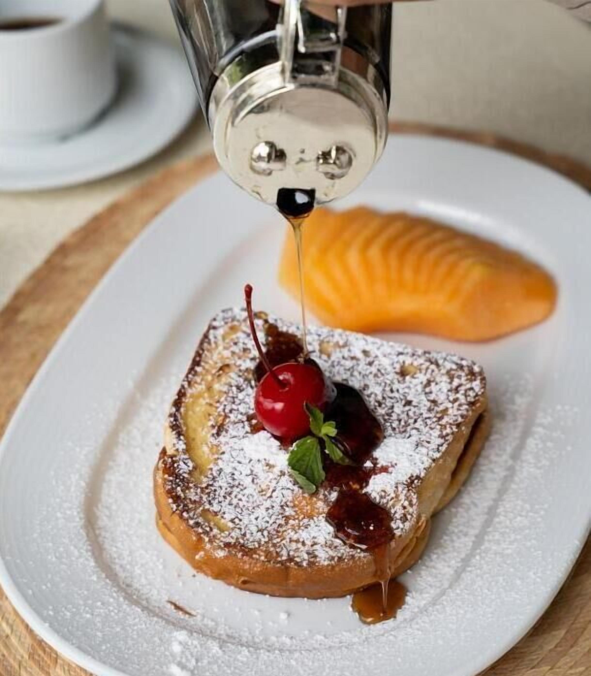 Un plato de postre con un pastel, una cereza, azúcar glas y jarabe siendo servido.