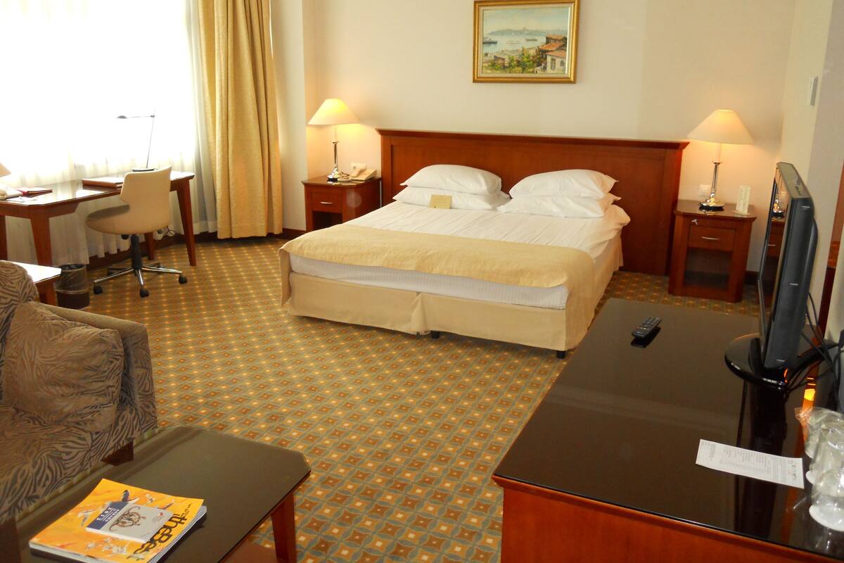 A hotel room with a bed, two bedside tables with lamps, a desk with a chair, a television, and a patterned carpet.
