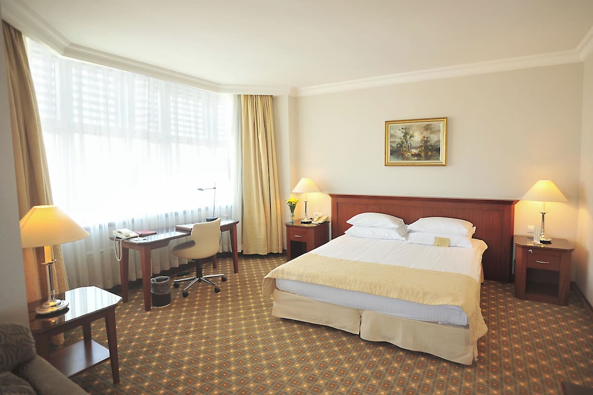 A hotel room with a large bed, two bedside tables with lamps, a desk with a telephone, a chair, and a window with curtains.