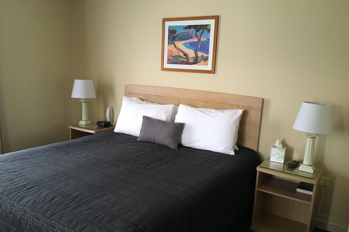 A neatly made bed with a dark bedspread and two pillows, flanked by nightstands with lamps and a tissue box.