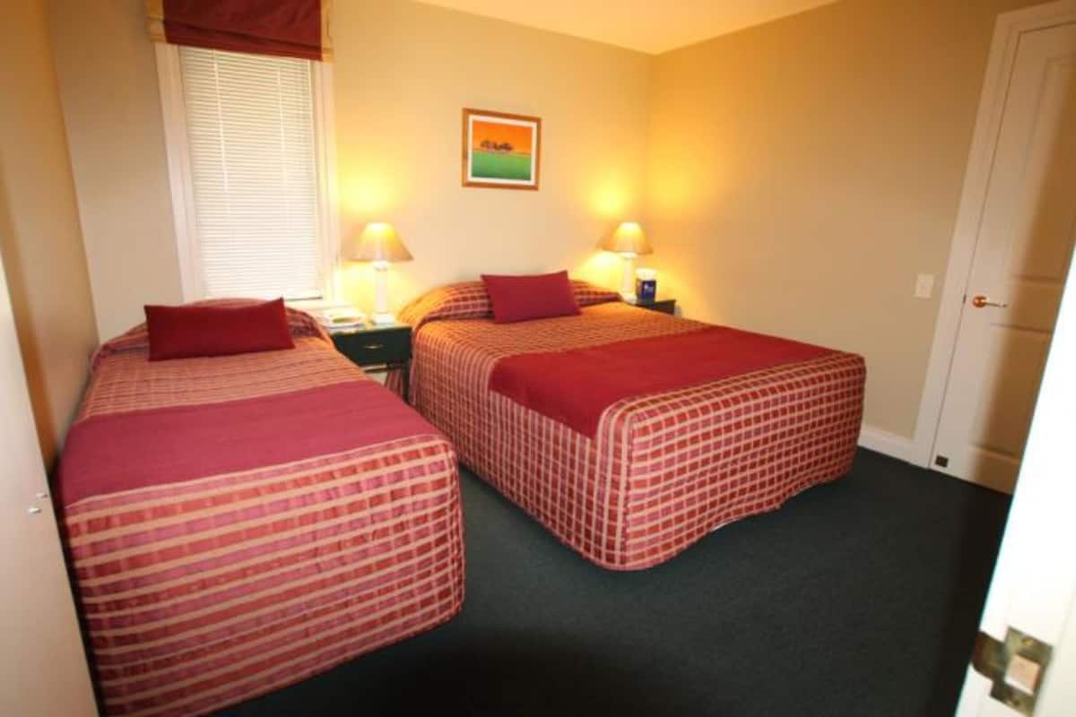 A room with two beds, each with a red checkered bedspread and a matching pillow. There are two bedside tables with lamps, a window with blinds, and a framed picture on the wall.