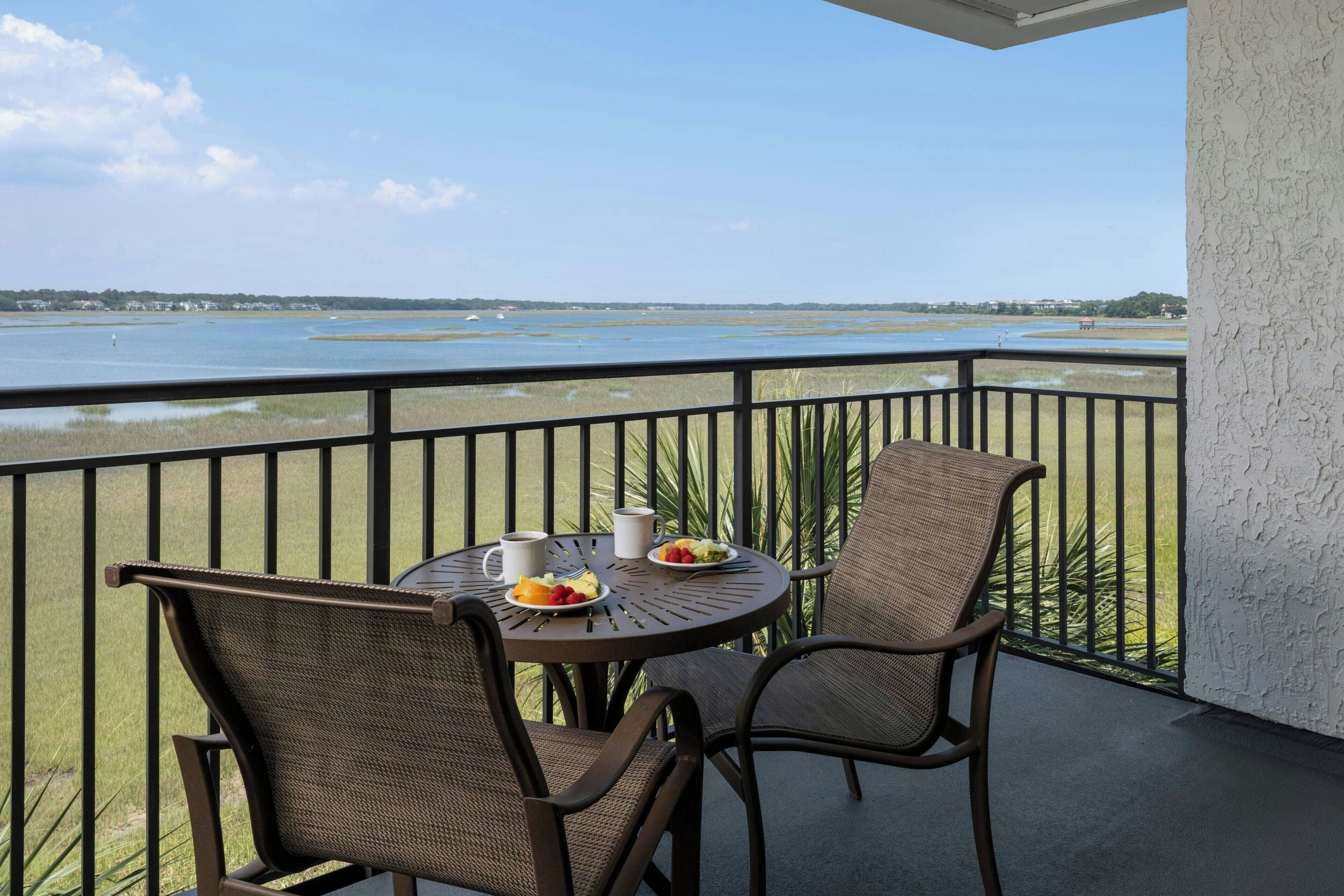 A balcony with two wicker chairs and a round table, overlooking a body of water with distant land and buildings.