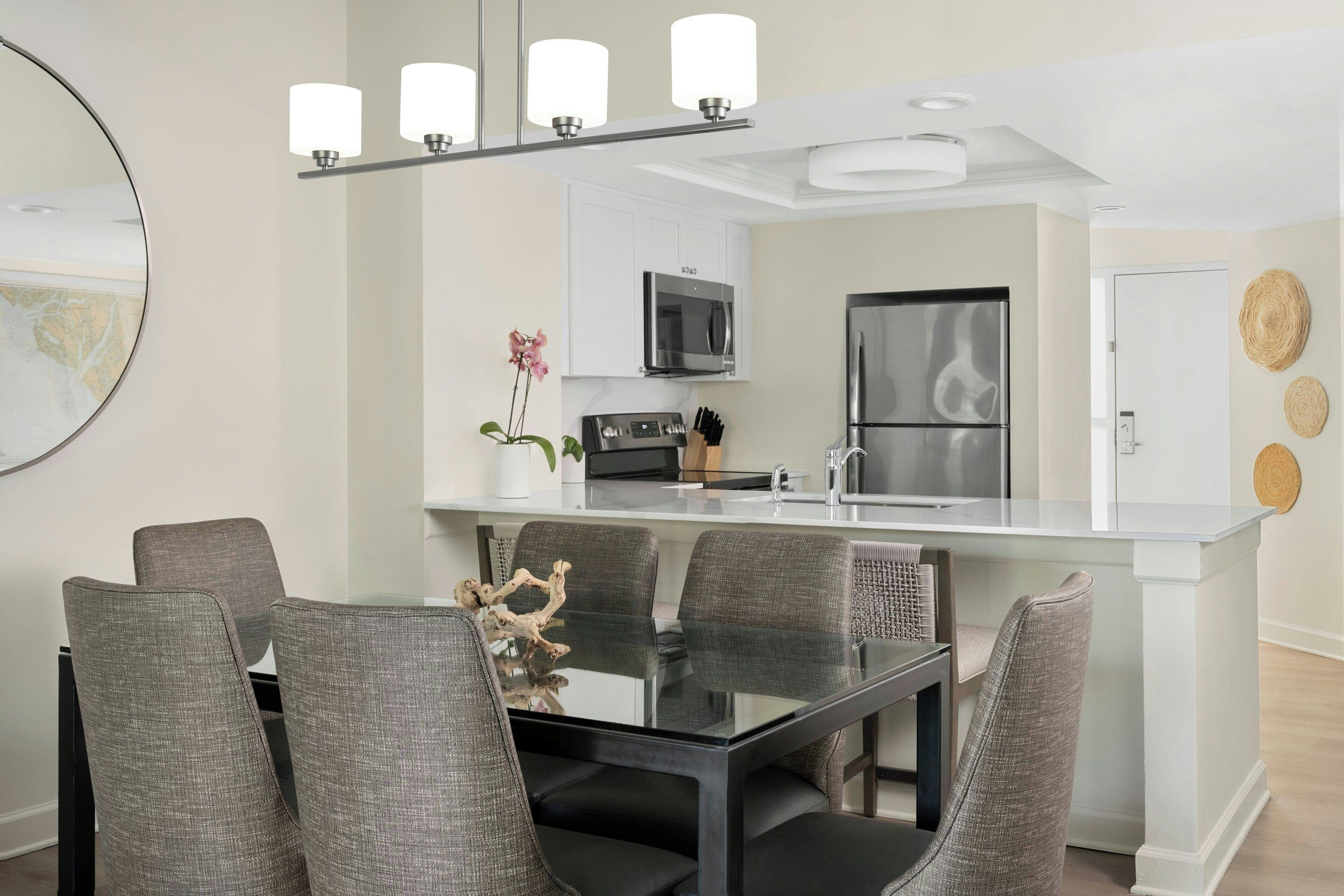 A modern kitchen with a central island, a dining area with a glass table and grey upholstered chairs, and a large mirror on the wall.