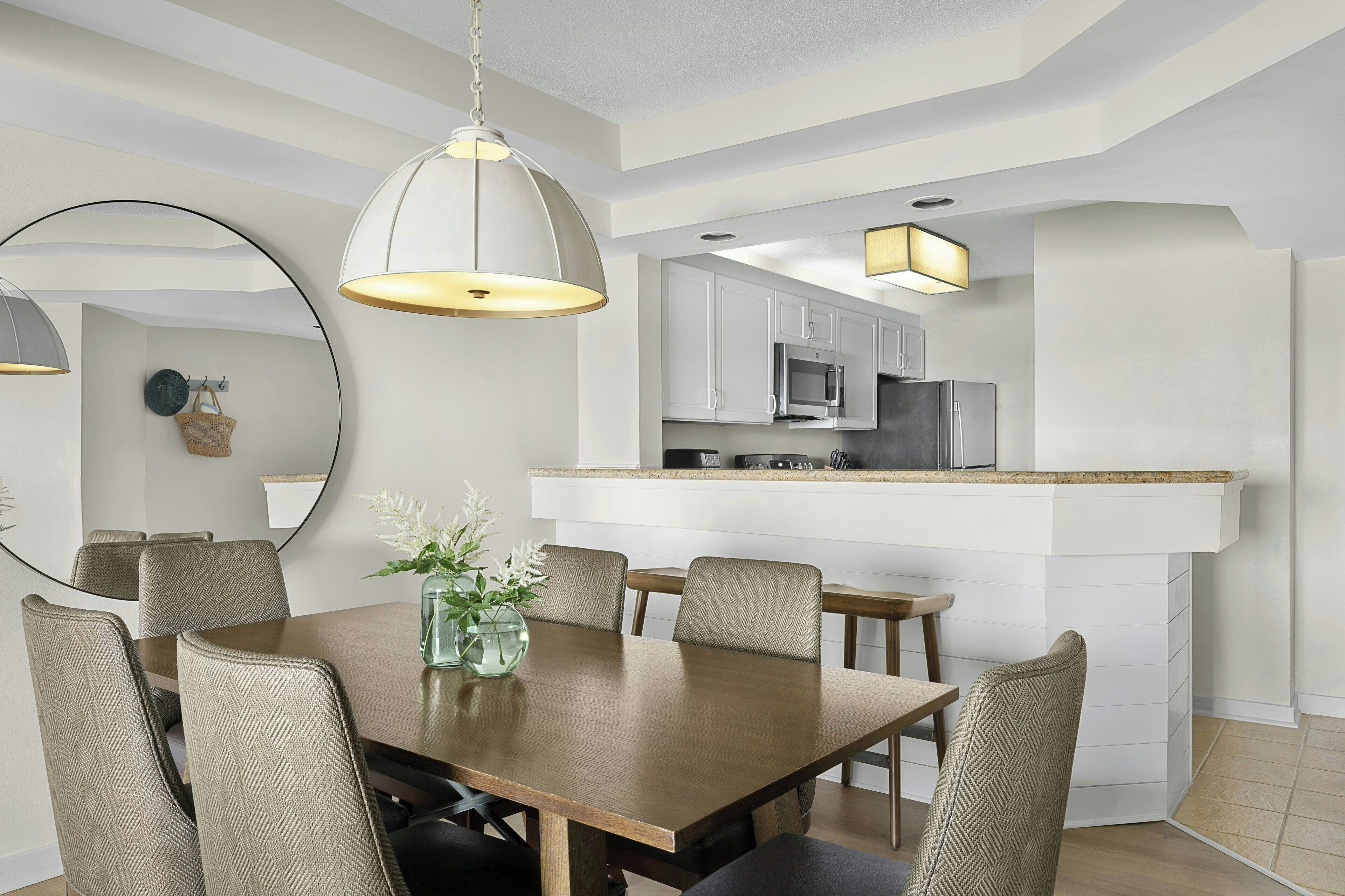 A modern dining area with a wooden table and chairs, a vase with flowers, and a kitchen in the background.