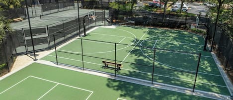 Una cancha de baloncesto al aire libre con un banco de madera, rodeada por una valla y árboles.