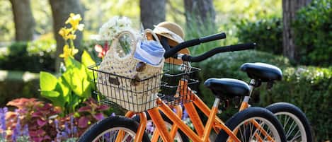 A bicycle with a basket containing a hat, a towel, and a small plant, parked in a garden with trees and flowers.