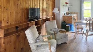 Una sala de estar acogedora con paneles de madera, un televisor de pantalla plana, dos sillones, una mesa de comedor con sillas y una zona de cocina con ventana.