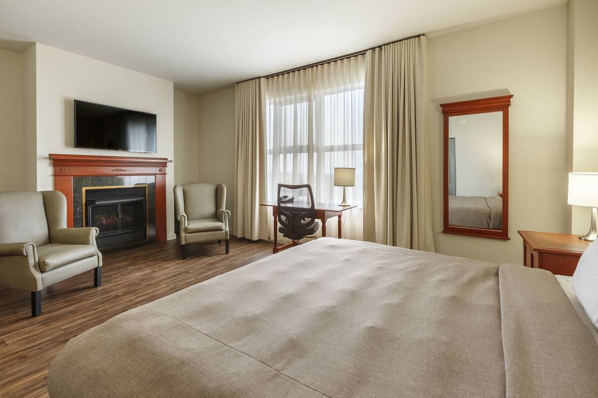 Ein Hotelzimmer mit einem großen Bett, zwei Sesseln, einem Kamin, einem Fernseher, einem Schreibtisch mit Stuhl, einem Spiegel und einem fenster mit Vorhängen.