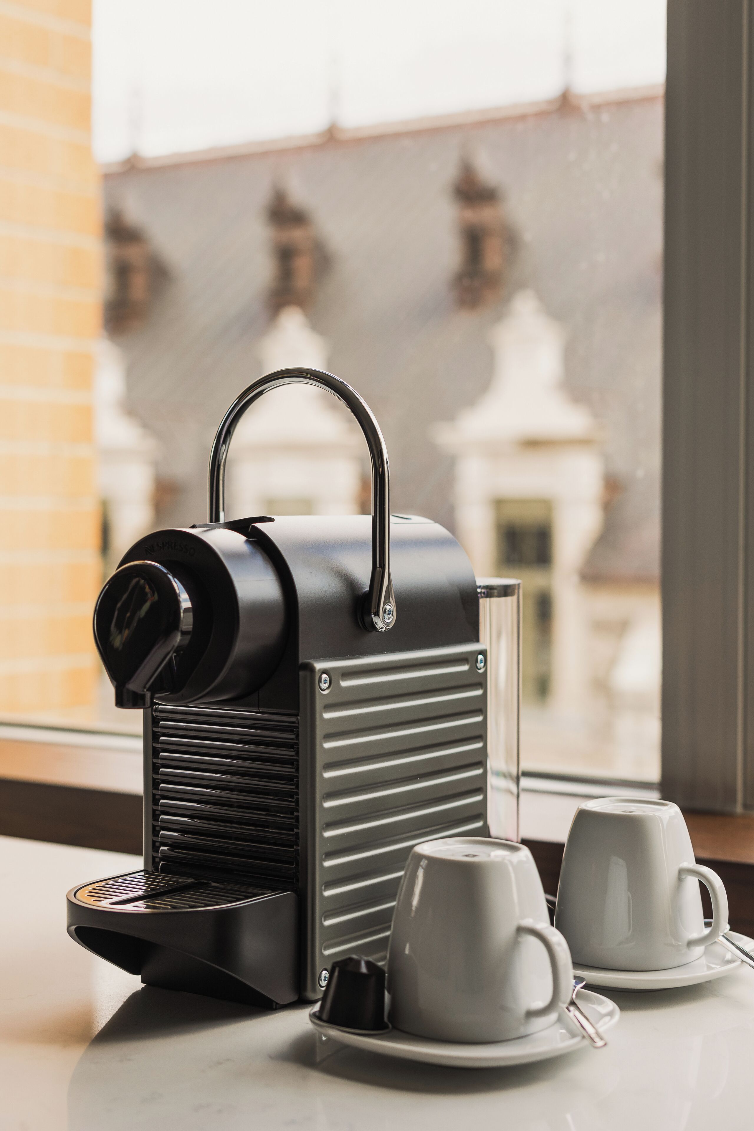 A coffee machine with two white cups on a table by a window.