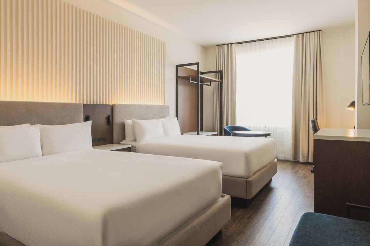 Ein Hotelzimmer mit zwei Betten, einem Schreibtisch, einem Stuhl und einem großen Fenster mit Vorhängen.